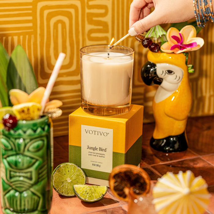 A hand lights a Votivo Jungle Bird 10 oz. Signature Candle, made with a soy blend, atop its box. Nearby are a green tiki drink, sliced limes, and a yellow parrot mug with a flower against a vibrant tropical backdrop.