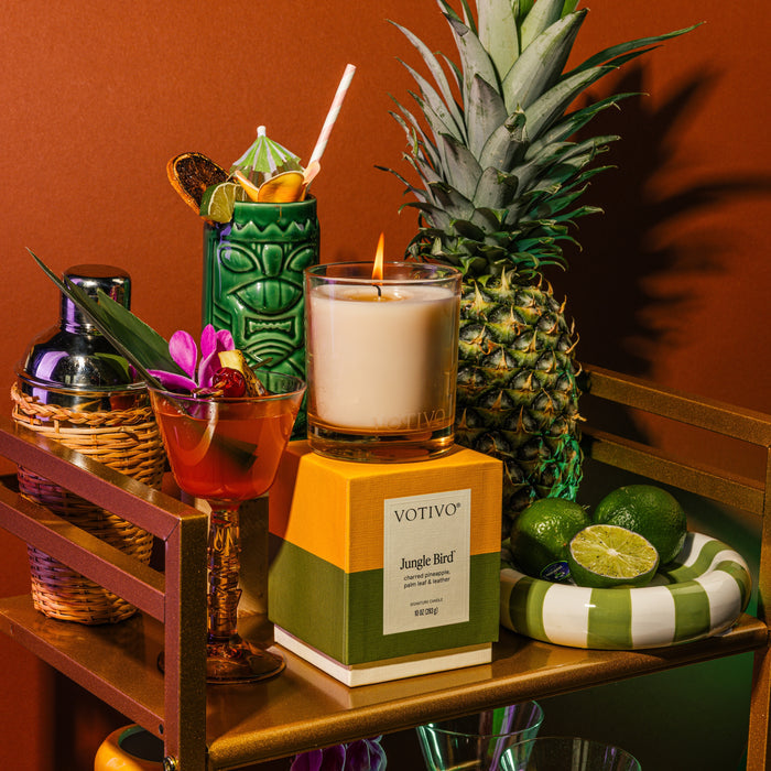 A gold bar cart displays a pineapple, Votivo’s Jungle Bird 10 oz. Signature Candle, a green tiki mug with a straw, a flower-garnished cocktail, fresh limes, and a striped dish set against a brown background.