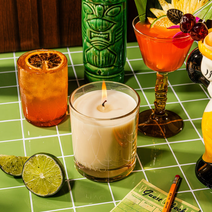 The Votivo Jungle Bird 10 oz. Signature Candle, crafted with a soy blend wax, burns in a glass on green tiles amid tropical cocktails, citrus garnishes, a halved lime, and a yellow guest check for a festive atmosphere.
