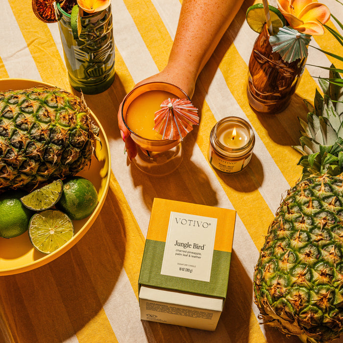 A tropical flat lay with pineapples, limes, a yellow-striped cloth, festive fruit- and flower-garnished drinks, and the Votivo Jungle Bird 10 oz. Signature Candle—a soy blend wax candle with a unique fragrance.