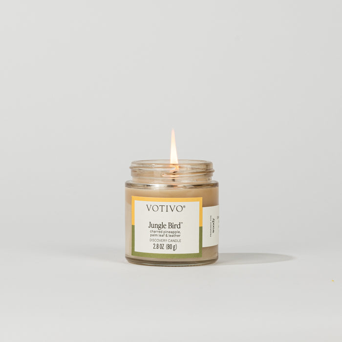A small Votivo Jungle Bird Discovery Candle burns with a single flame against a white background. This soy blend candle has a white and green label and contains 2.8 oz (80g) of long-lasting fragrance.