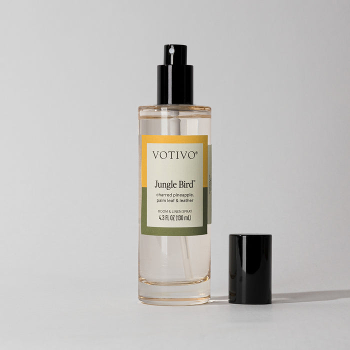 A clear glass bottle of Jungle Bird Room & Linen Spray with a black spray nozzle. The label shows the product name and fragrance description.