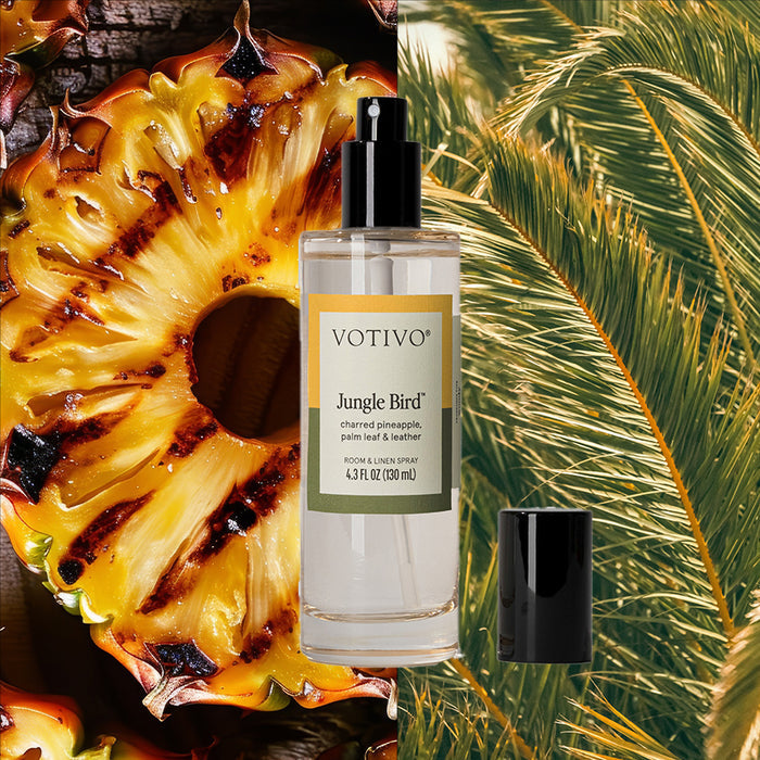 A Votivo Jungle Bird Room & Linen Spray stands with its black cap beside it, set against a backdrop of charred pineapple and palm leaves, infusing fabrics and rooms with the bold Jungle Bird fragrance.