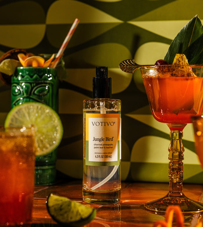 A bottle of Votivo Jungle Bird Room & Linen Spray rests on a table with colorful cocktails, lime wedges, and garnishes, set against a retro green and yellow patterned background.