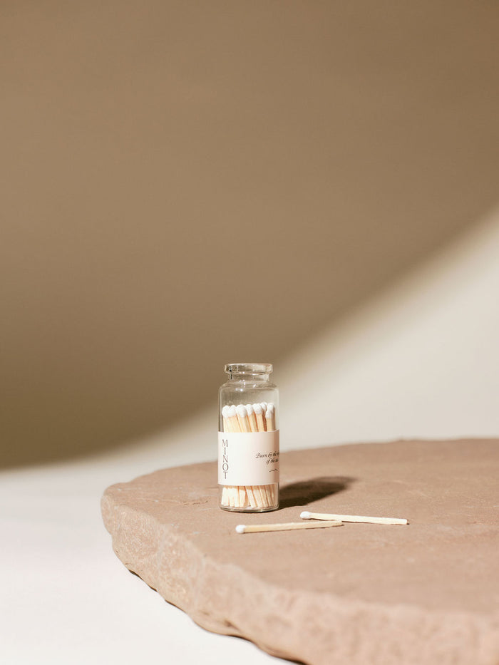 A small glass jar of matches with a white "MINOT" label sits on a textured stone surface. Two matches lie beside the jar. The background is softly lit with shadows.