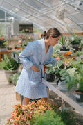 Woman in a blue patterned robe examines potted plants on a metal shelf in a greenhouse. Various green plants and flowers surround her in the bright setting.