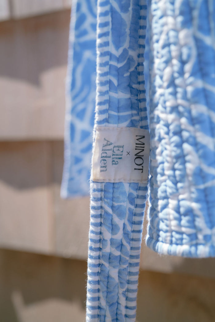 Close-up of a light blue quilted fabric with a white label reading "MINOT x Ella Alden." The fabric features a subtle coastal pattern, hanging against a blurred background.