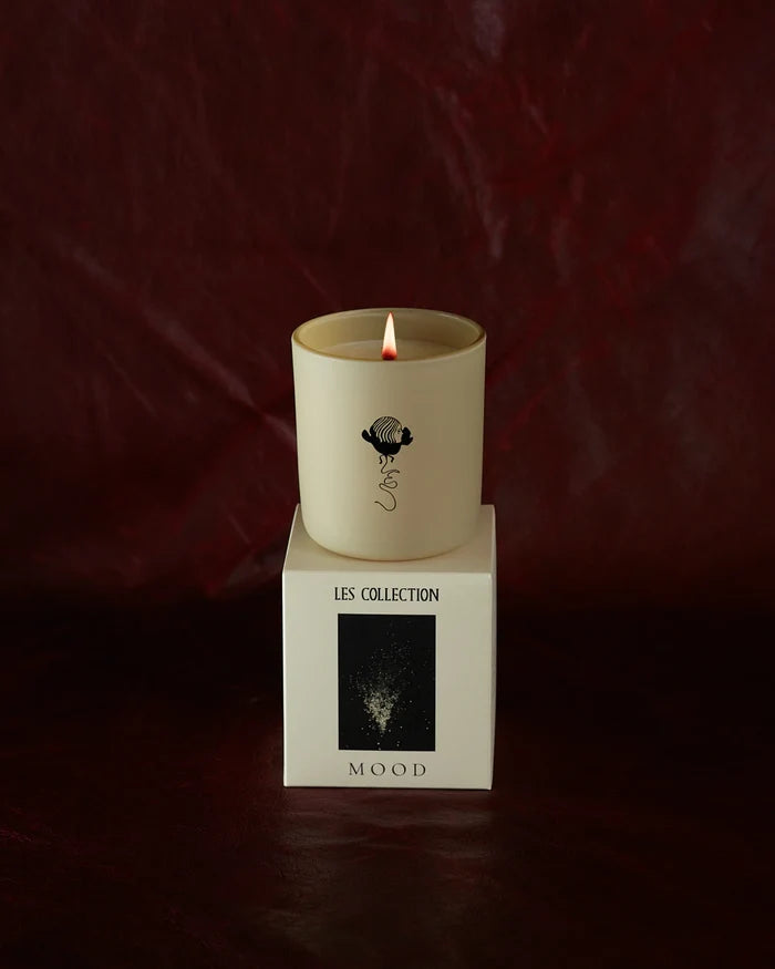 A lit LES Collection Mood candle in a glass jar sits on a white box labeled "LES Collection Mood" against a dark red background.