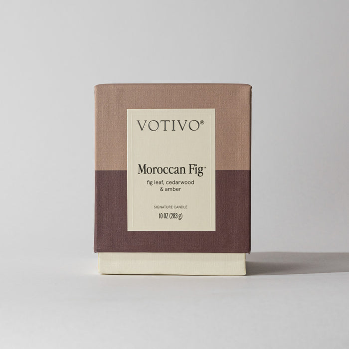 A Votivo Moroccan Fig 10 oz. Signature Candle in a minimalist beige, taupe, and brown box features notes of fig leaf, cedarwood, and amber. Made with soy blend wax, the candle is shown against a plain background.