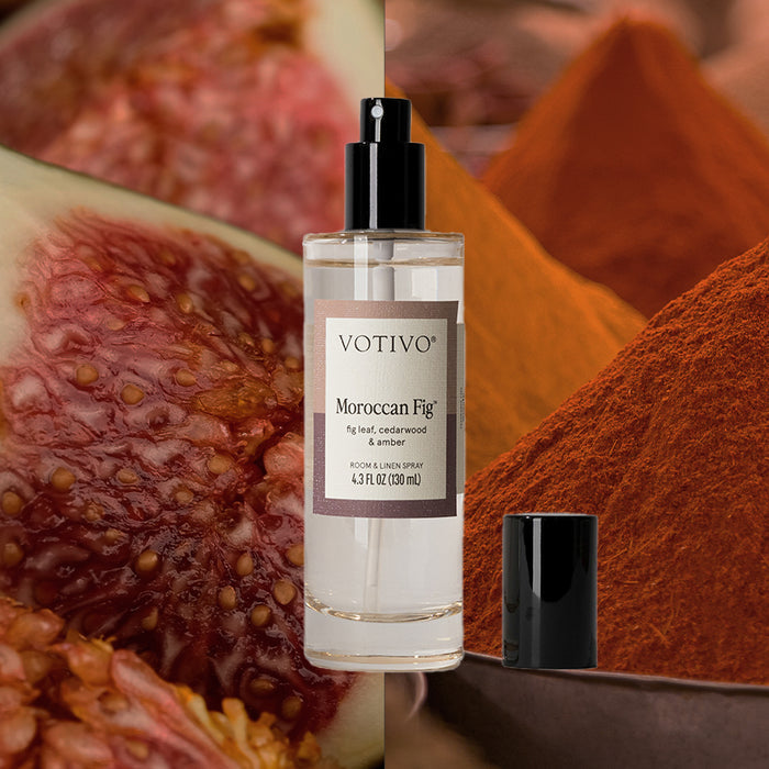 A Votivo Moroccan Fig Room & Linen Spray stands upright with its cap off, set before close-up figs and piles of orange and red spices.