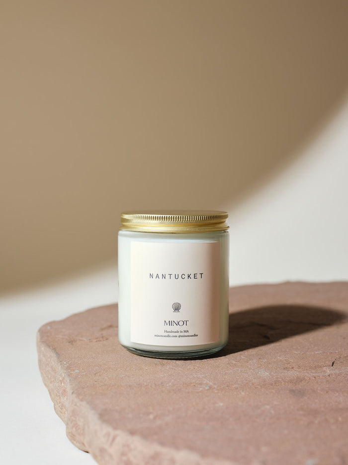 A minimalist soy candle jar with a gold lid labeled "Nantucket" and "MINOT" sits on a textured stone surface against a neutral background.