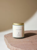 A minimalist soy candle jar with a gold lid labeled "Nantucket" and "MINOT" sits on a textured stone surface against a neutral background.