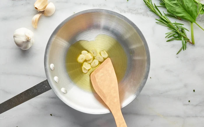 A stainless steel saucepan with a wooden spoon on a light marble surface, containing oil and garlic cloves, with garlic, basil, and rosemary arranged around it.