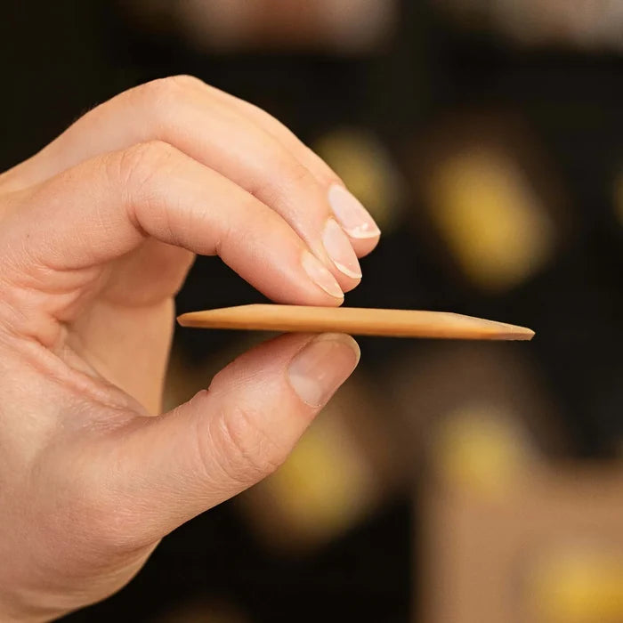 Side view of pan scraper held between thumb and forefinger showcasing thin, flat beveled edge and wood grain against blurred background