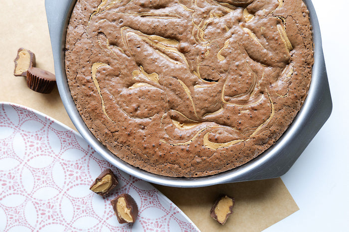 Top view of 9-inch round brownie pan showcasing peanut-butter swirl brownie and side handles on a white surface with patterned plate and halved peanut butter cups