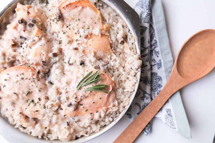 Skillet of creamy mushroom chicken and rice topped with rosemary and cracked black pepper, wooden spoon to the right.