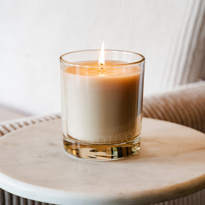 The Votivo Pink Mimosa 10 oz. Signature Candle, crafted with a soy blend wax and set in a clear glass holder, sits on a round white marble table, its gentle glow adding subtle elegance to any space against a soft textured backdrop.