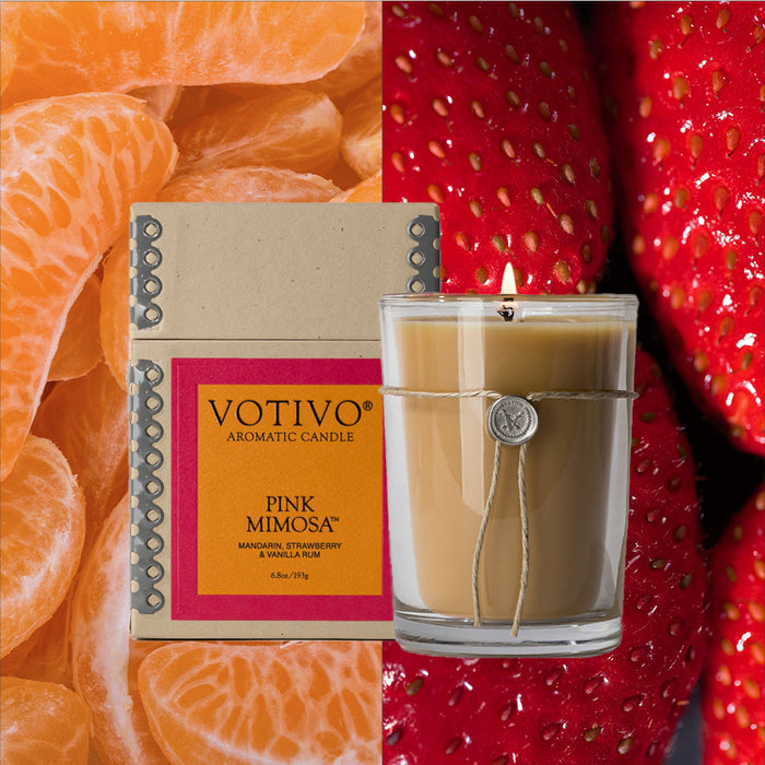 A Votivo Pink Mimosa 6.8 oz. Aromatic Candle and its box sit among mandarin orange segments and ripe strawberries. Made with a soy blend wax, the candle is tied with string and topped by a signature wax seal.