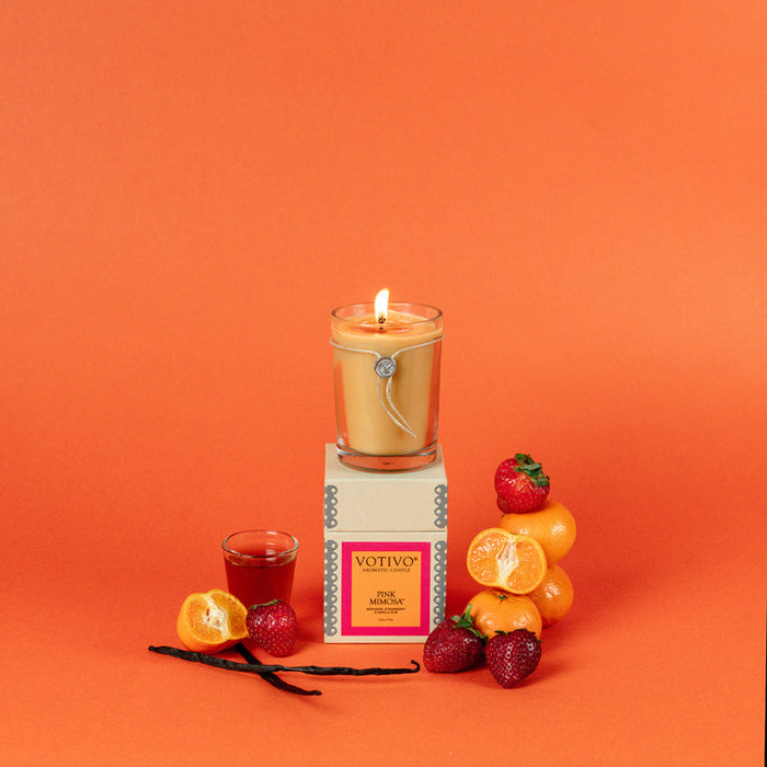 A Votivo Pink Mimosa 6.8 oz. Aromatic Candle sits on its box, surrounded by strawberries, tangerines, vanilla beans, and a glass of red liquid. Set against an orange background. Made with soy blend wax for a clean burn.