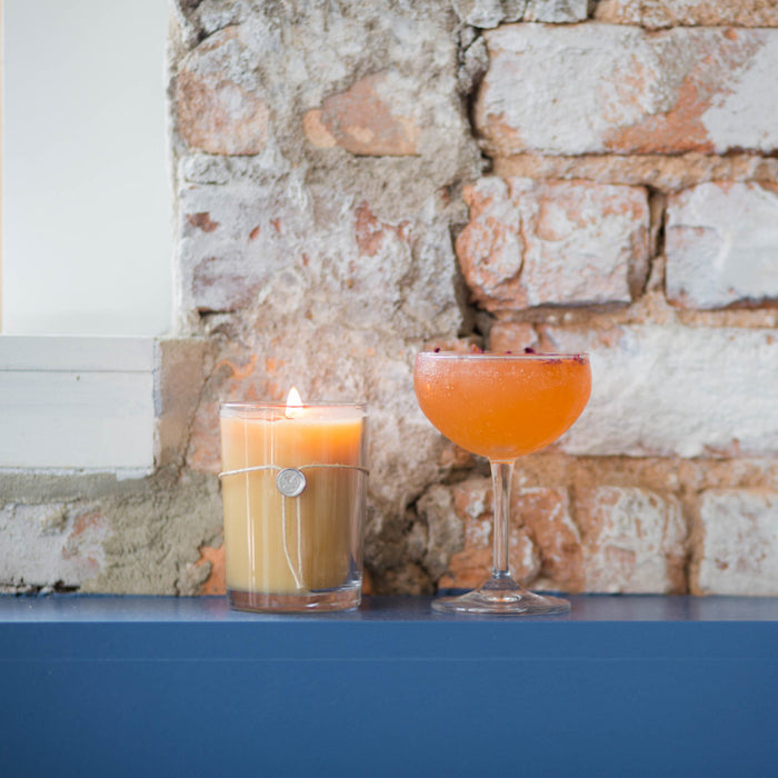 The Votivo Pink Mimosa 6.8 oz. Aromatic Candle in a glass jar and an orange cocktail rest on a blue ledge before a rustic brick wall, setting the scene for relaxation.