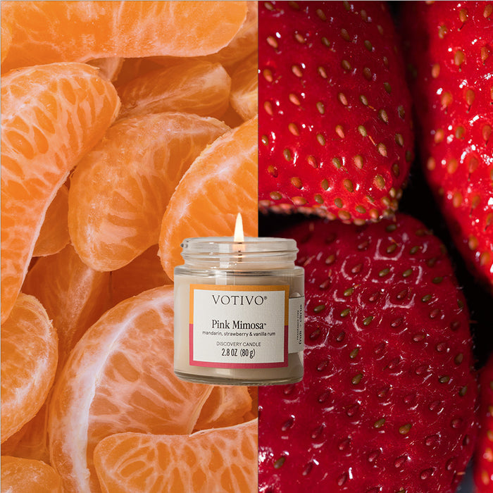 A Votivo Pink Mimosa Discovery Candle is lit, set before a split background with tangerine segments on the left and strawberries on the right.