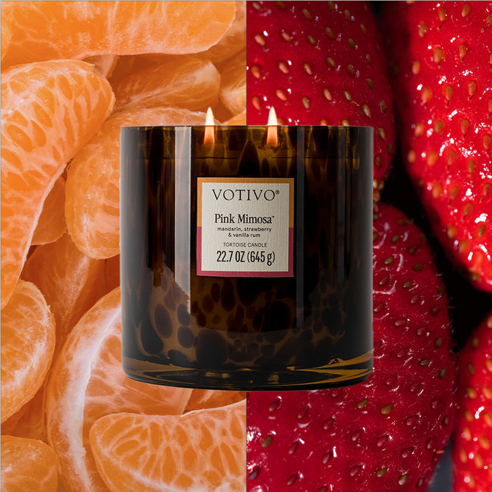 A Votivo Pink Mimosa Tortoise Candle with three lit wicks sits in a dark glass jar, centered over a split background of orange tangerine segments on the left and red strawberries on the right.