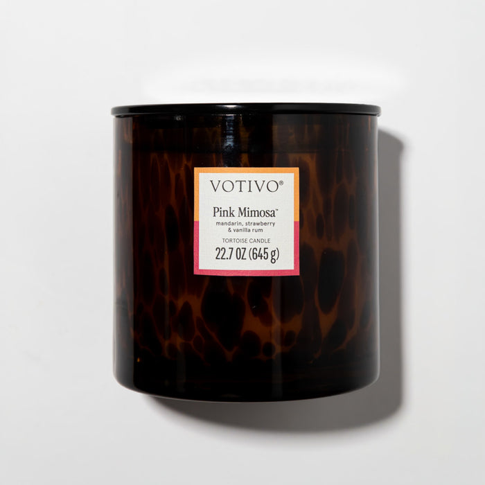 The Votivo Pink Mimosa Tortoise Candle is a brown, tortoiseshell-patterned soy blend with 22.7 oz (645 g) of wax and notes of mandarin, strawberry, and vanilla rum, shown resting on a white surface.