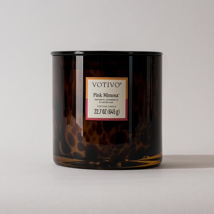 A Votivo Pink Mimosa Tortoise Candle in a dark, mottled glass jar displays the fragrance name and weight (22.7 oz/645 g) on its label, set against a plain light background.