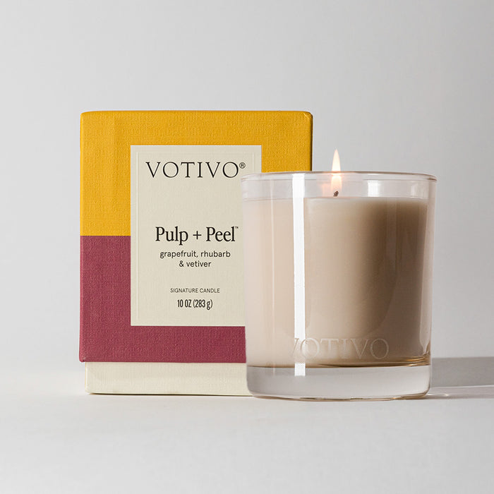 A lit Votivo Pulp + Peel 10 oz. Signature Candle in clear glass sits before its yellow and burgundy box, featuring notes of grapefruit, rhubarb, and vetiver. Crafted from a soy blend wax for a unique scent experience.