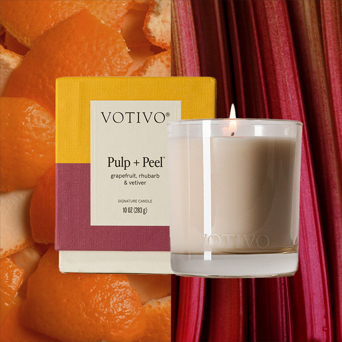 A lit Votivo Pulp + Peel 10 oz. Signature Candle in a clear glass holder stands before its colorful box, with grapefruit peels to the left and rhubarb stalks to the right, releasing its unique fragrance.