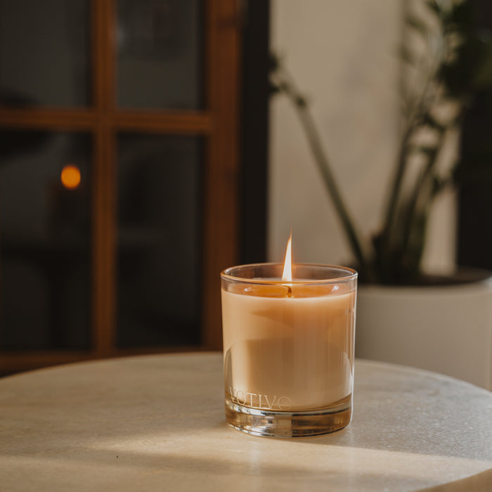 A lit Votivo Pulp + Peel 10 oz. Signature Candle in a glass holder rests on a round light table, with a plant and blurred window softly illuminating the background.