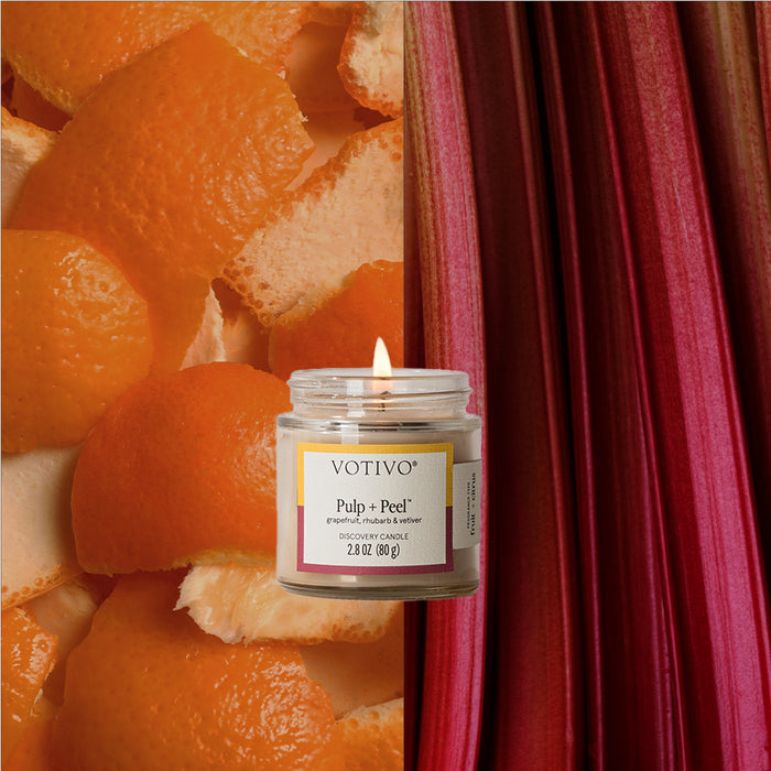 A burning Votivo Pulp + Peel Discovery Candle sits against a backdrop with orange peels on the left and red rhubarb stalks on the right.
