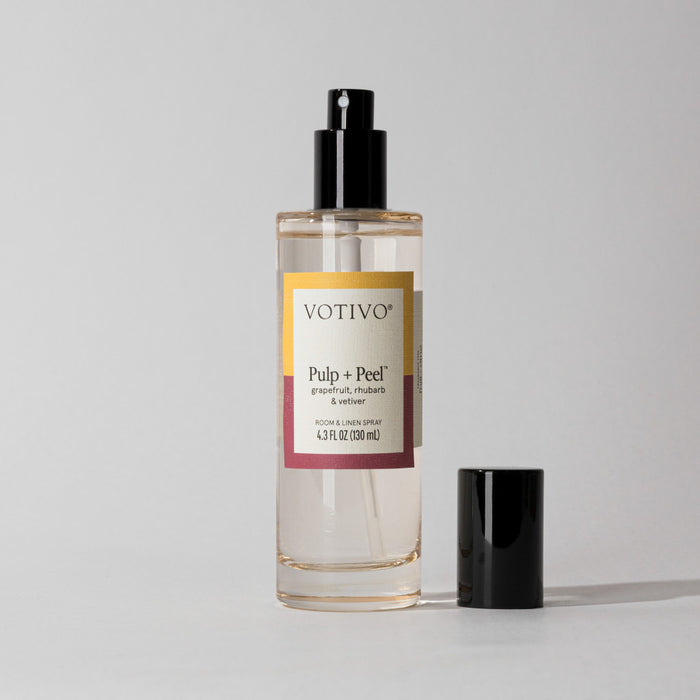 A clear glass bottle of Votivo Pulp + Peel Room & Linen Spray with a black nozzle and cap sits beside it. The label showcases its citrus scent—grapefruit, rhubarb, vetiver—and notes its fabric safe. Set against a plain light background.