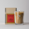 A lit Votivo Red Currant 6.8 oz. Aromatic Candle glows in a clear glass jar, tied with string and seal, placed before its decorative box on a light gray background.