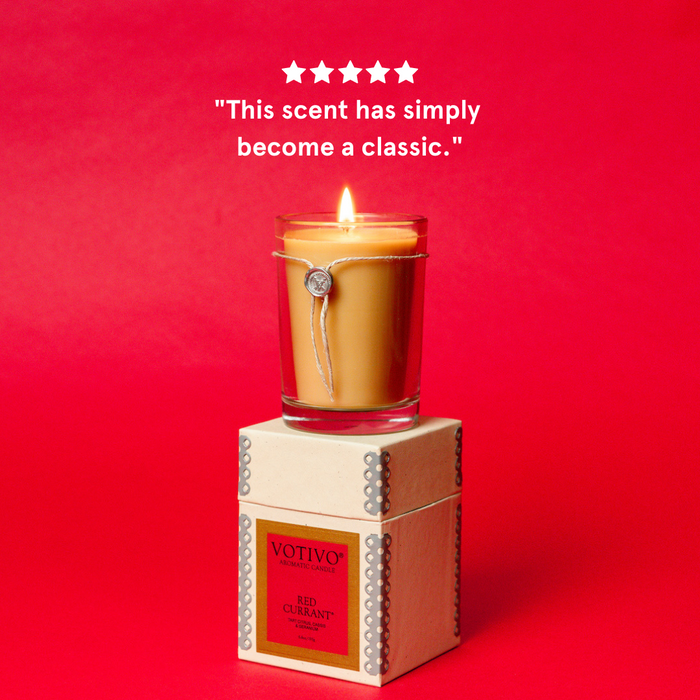 A Votivo Red Currant 6.8 oz. Aromatic Candle burns atop its box against a red backdrop. Above, five stars and the quote: “This scent has simply become a classic” highlight the signature fragrance’s acclaim.