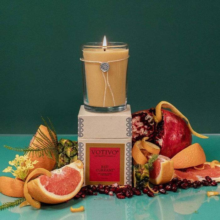 The Votivo Red Currant 6.8 oz. Aromatic Candle sits atop its box, surrounded by grapefruit slices, pomegranate, orange peels, and green leaves on a teal background. Its vibrant red currant scent fills the air.