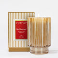 A Votivo Red Currant Holiday Candle, a luxurious soy blend in a ribbed glass holder, stands before its striped beige and gold box with a red label on a plain white background.