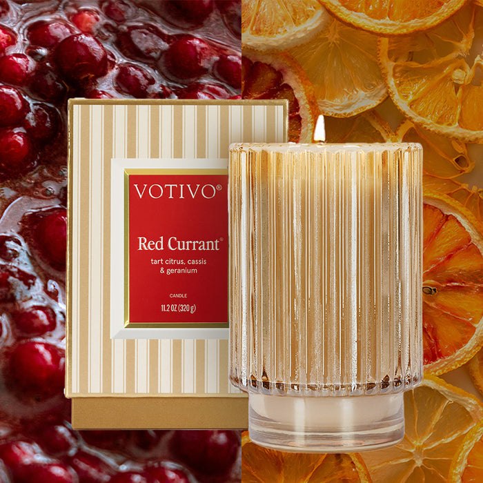 A Votivo Red Currant Holiday Candle in a ribbed glass jar sits beside its striped box, with red currants on the left and dried orange slices on the right.