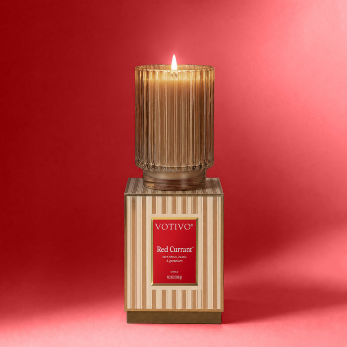A lit Votivo Red Currant Holiday Candle, made with soy blend wax in a ribbed glass container, sits atop its decorative striped box with a red label, set against a vibrant red background.