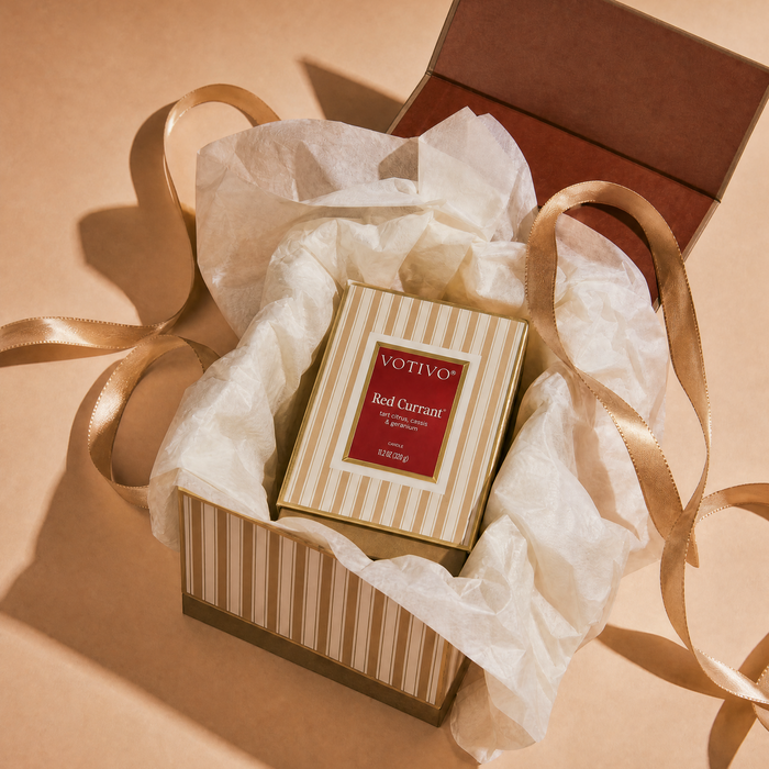 A Red Currant Holiday Boxed Candle in a striped gift box with tissue paper and ribbon on a tan background