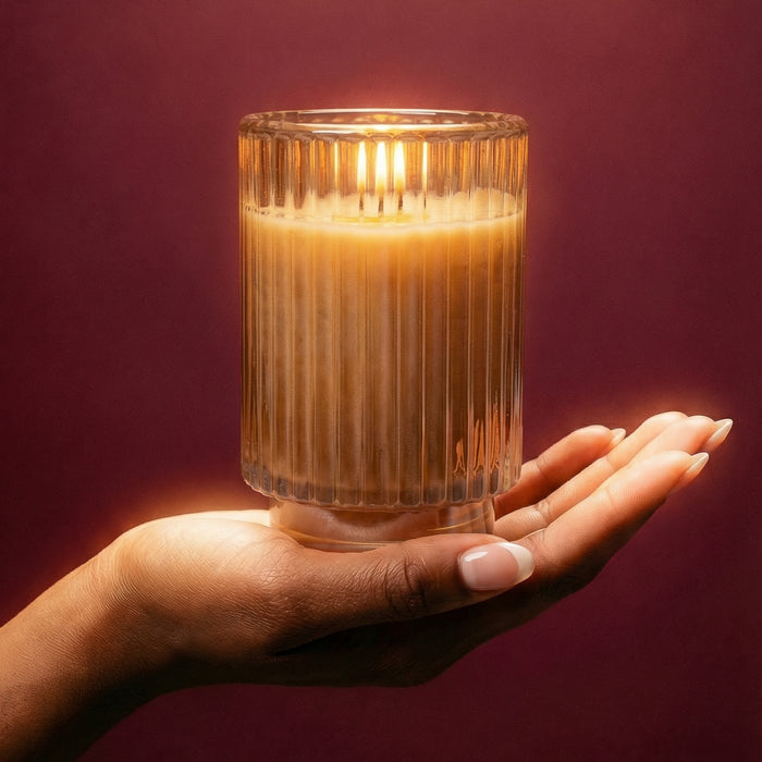A hand with manicured nails gently holds a lit Votivo Icy Blue Pine Holiday Candle made from soy blend wax in a ribbed glass jar against a warm burgundy background, casting a soft glow.