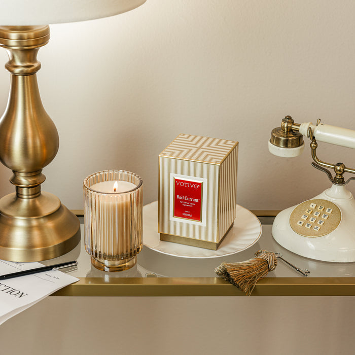 A lit Votivo Red Currant Holiday Candle, its box nearby, is arranged neatly on a glass-topped table alongside a vintage phone and a gold lamp.