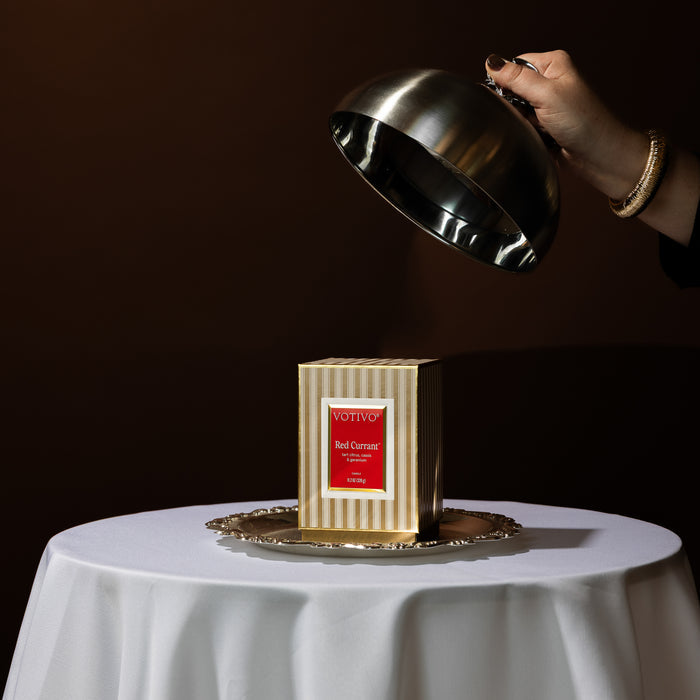 A hand lifts a silver cloche to unveil the Votivo Red Currant Holiday Candle, presented in its striped box on a round silver tray atop a white tablecloth, set against a dark brown background.
