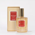 A Votivo Red Currant Holiday Room & Linen Spray with a gold cap stands before its matching gold and white striped box, both adorned with red labels and white text highlighting the signature red currant scent.