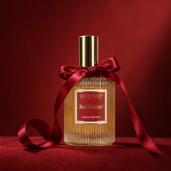 A bottle of Votivo Red Currant room and linen spray, featuring a ribbed clear glass body with amber liquid, a gold cap, and a red satin ribbon bow tied around the neck, set against a dark red background.