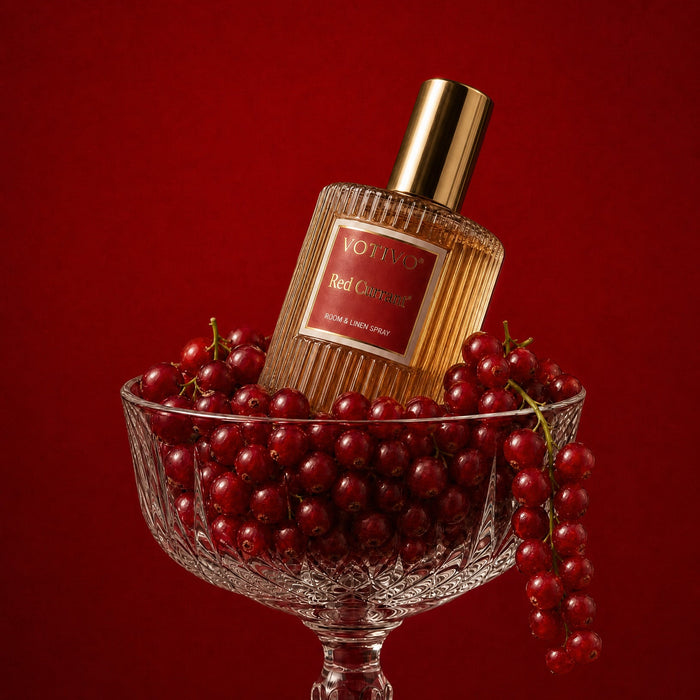 A bottle of Red Currant Room & Linen spray with a gold cap, nestled in a glass bowl filled with red cranberries against a solid dark red background.