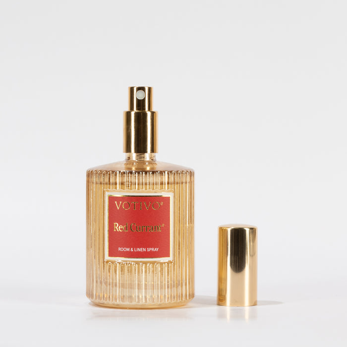A ribbed glass bottle of Votivo Red Currant Holiday Room & Linen Spray with a gold nozzle and cap, displayed on a white background. The cap sits beside the bottle, emphasizing its signature red currant scent.