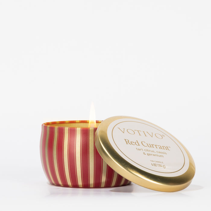 A lit Votivo Red Currant Holiday Tin Candle in a red and gold striped tin with its lid, reading "VOTIVO Red Currant," leaning nearby. The scent features tart citrus, cassis, and geranium. Background is white.