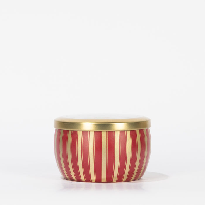 The Votivo Red Currant Holiday Tin Candle features vertical red and gold stripes with a matching gold lid, crafted from soy blend wax, and is shown against a plain white background.