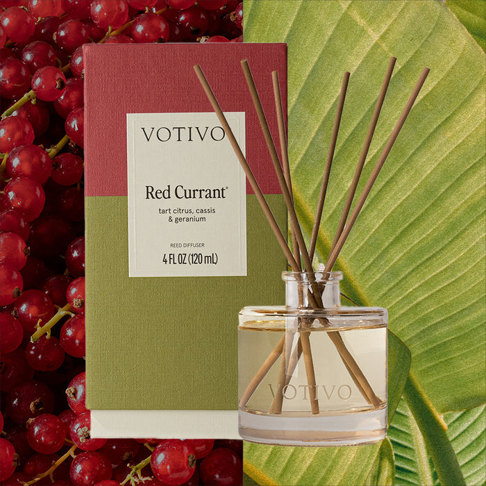 A Votivo Red Currant Reed Diffuser is displayed in front of its box, surrounded by red currants and green leaves. The clear glass bottle holds premium fragrance and includes several brown fiber reeds inside.